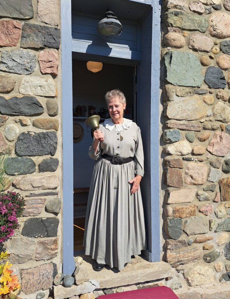 Clara Michaluk, dressed in period costume for October’s Fall Family Festival, rings the teacher’s bell as she stands in the schoolhouse doorway at the Wooden Old Stone School near Onsted.