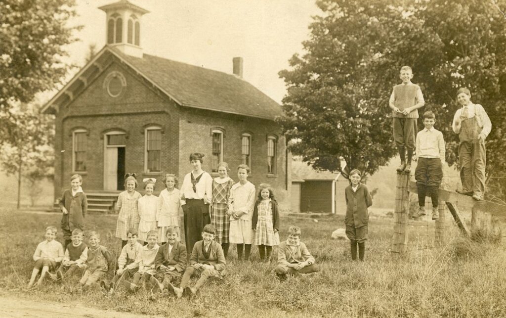 The Maynard school on Hamilton Highway west of Adrian in 1919. Sitting: Arthur Snyder, Lewellyn Garrison, Harley Crist, George Maynard, Richard Brittain, Pierson Rathbun, Leroy Younglove, and Charles Rathbun. Standing: Olen Iffland, Elizabeth Brittain, Mary Alice Younglove, Gladys Crist, Ida Woodruff (the teacher, 18 years old), Marcella Rathbun, Grace Prichard, Marjorie Cuer, Therol Brittain, and on the fence: Carson Garrison, Llewellyn (Bill) Crist, and George Rathbun. (Melvin Snyder photo collection)