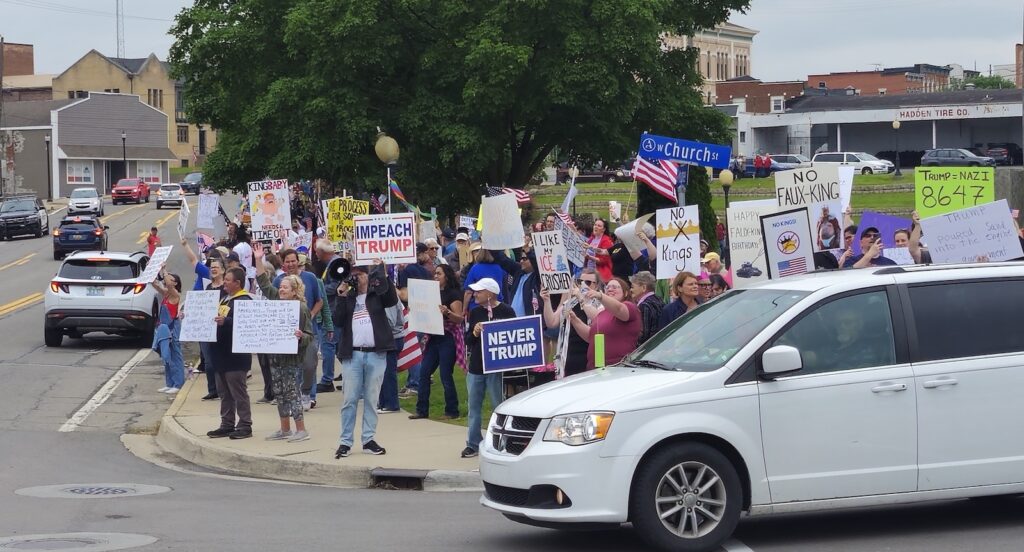Opponents of the Trump administration line the edge of Comstock Park at the corner of Maumee and Church streets, with many signs bearing variations on the phrase "No kings" and one depicting Trump as an infant with the words "King Baby needs a time out."