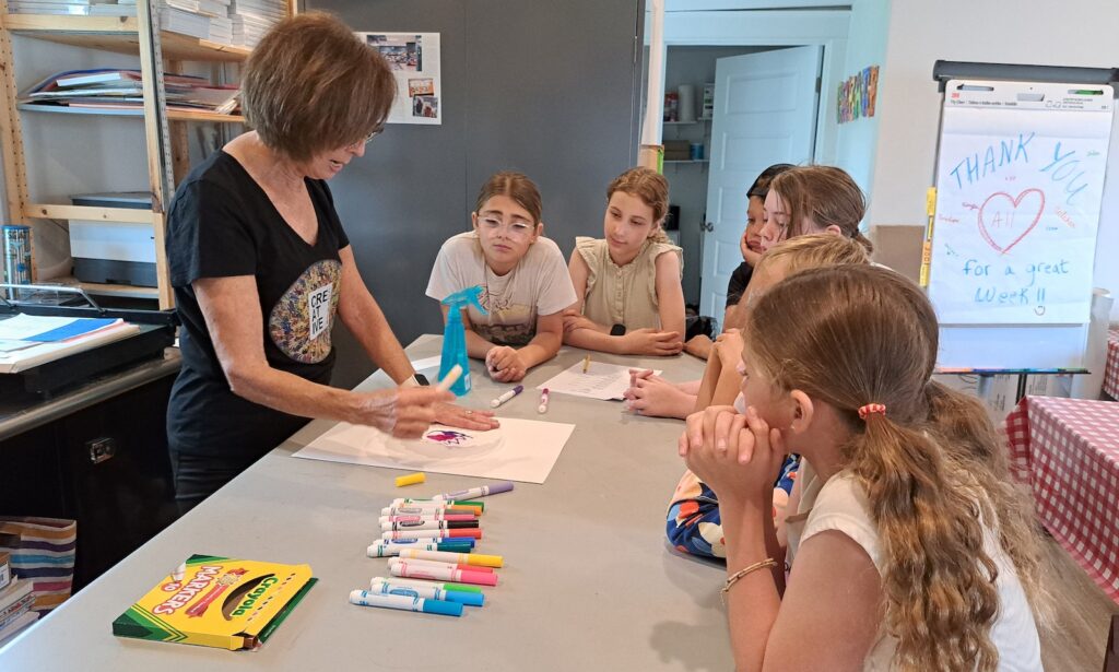 Jean Lash, the founder of Manitou Beach Creative Corner, shows students how to use a coffee filter, markers, and water to make a unique picture during the first arts camp of the summer on June 20. (Photo by Arlene Bachanov)