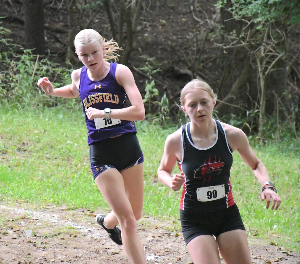Blissfield’s Mollie Yeager and Clinton’s Ella Claus go downhill and round a corner midway through the course at the Lenawee County cross country preview meet hosted by Tecumseh.
